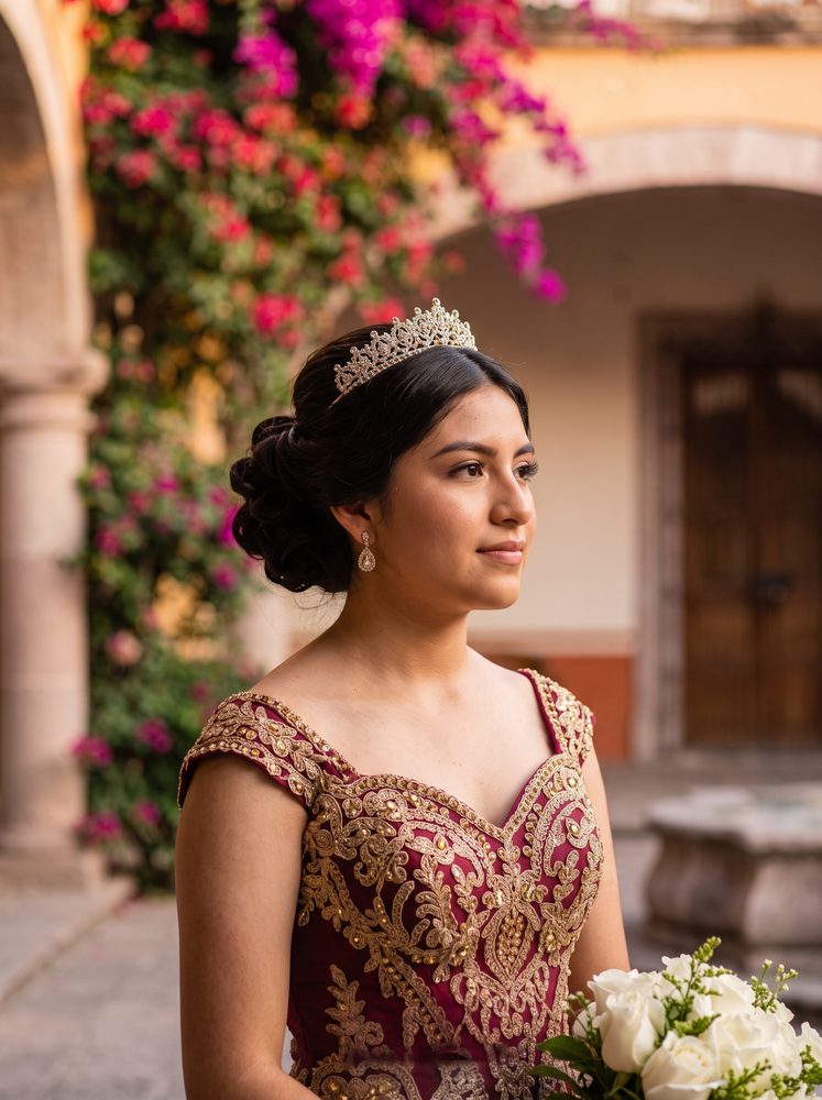 Valeria con tiara y vestido de XV años color vino bordado en dorado, bajo las buganvilias de un patio de cantera